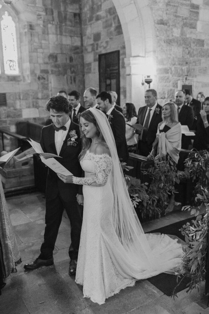 A bride and groom stand together, holding papers, inside a stone church during their wedding ceremony. Captured by a Creative Wedding Photographer, the bride wears a long lace gown and veil; guests stand behind them, watching attentively.