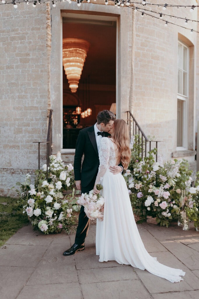 A bride and groom kiss outside a stone building adorned with flowers, captured perfectly by a creative wedding photographer. The bride’s white dress and bouquet and the groom’s black tuxedo glow under string lights and a sparkling chandelier.