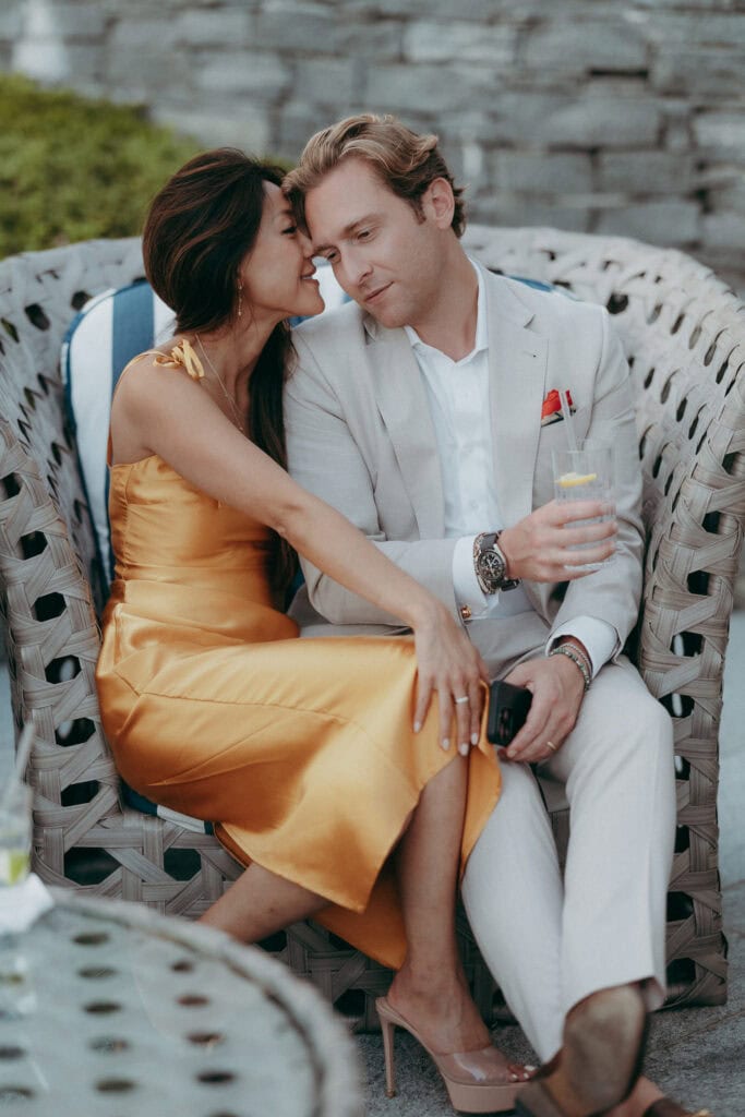 A woman in a gold satin dress and a man in a light suit sit closely on a woven outdoor chair. Captured by a Creative Wedding Photographer, she leans in tenderly while he holds a drink, both appearing relaxed and affectionate.