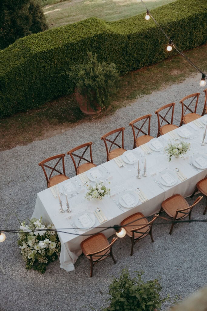 An elegant outdoor dining table is set with white linens, plates, and floral centerpieces—perfectly styled for a creative wedding photographer—surrounded by wooden chairs on gravel, with string lights and manicured hedges in the background.