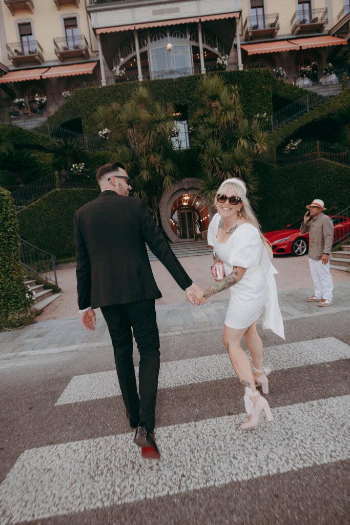 A couple holding hands crosses a street towards a lush, grand hotel. Captured by a creative wedding photographer, the woman in a white dress smiles back in sunglasses, while the man in a dark suit looks ahead. A red car and another person are visible behind them.