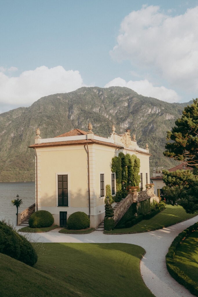 A yellow villa with tall windows and manicured gardens stands by a lake, with green mountains rising in the background—an idyllic setting perfect for a creative wedding photographer to capture unforgettable moments under a partly cloudy sky.
